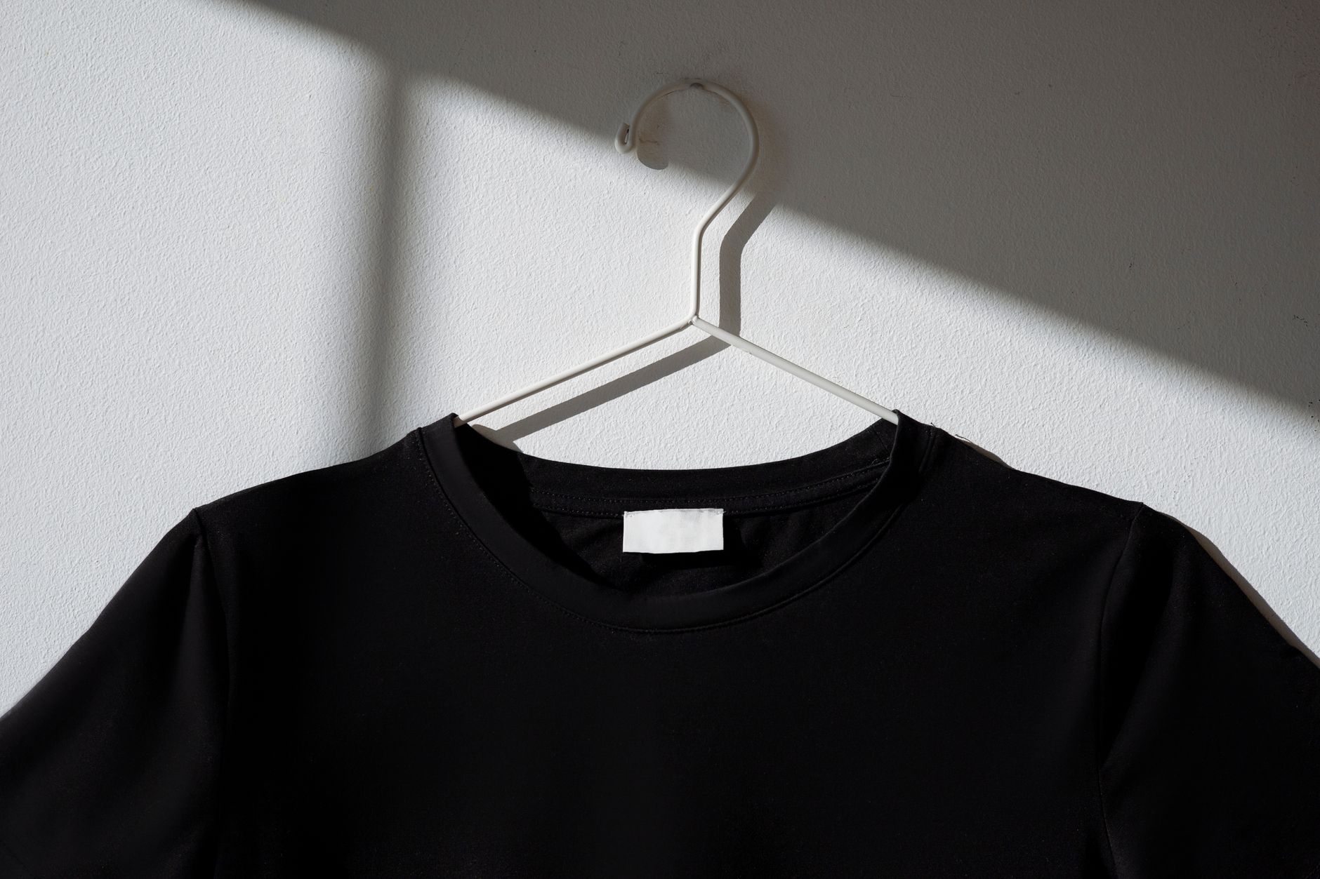 Black t-shirt hanging on a white hanger against a plain wall with a dramatic shadow effect.